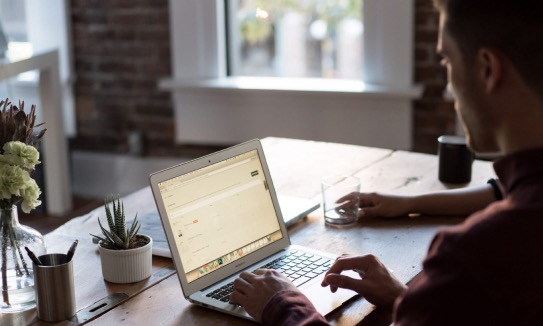 Writer typing on a laptop at a wooden desk beside a bright loft window, plants and a glass of water nearby